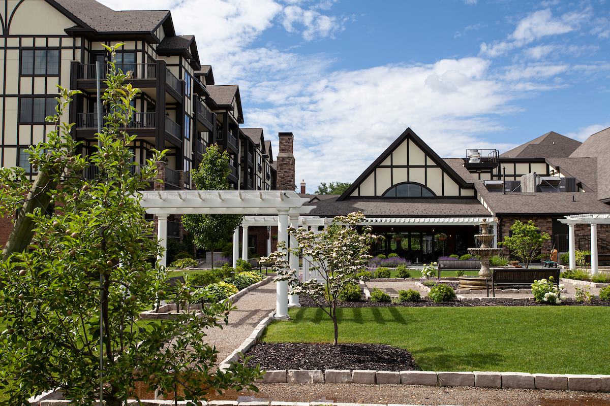 Paved pathways through a garden with white trellises, black benches, and a small stone fountain. Apartments with balconies in the background.uilding in the background.